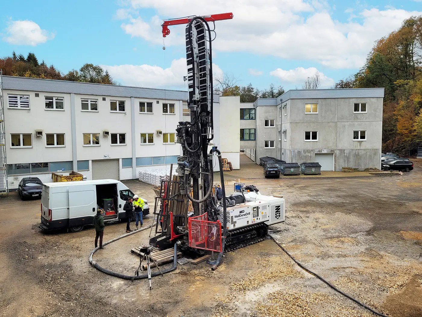 Machine de forage géothermique en action sur un chantier en Belgique, avec des techniciens travaillant à côté de véhicules et d'un bâtiment administratif.
